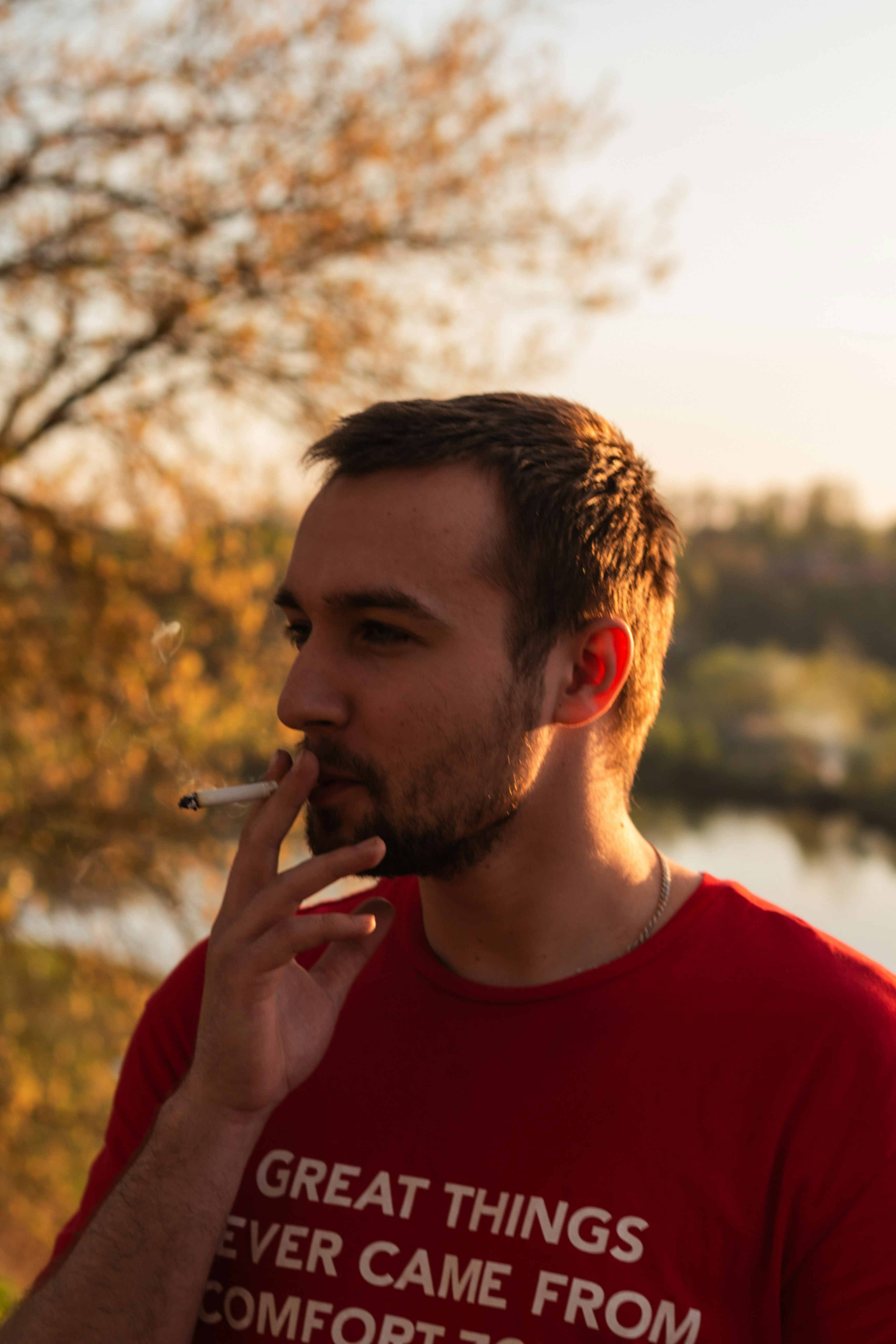 man standing smoking cigarette near body of water and trees